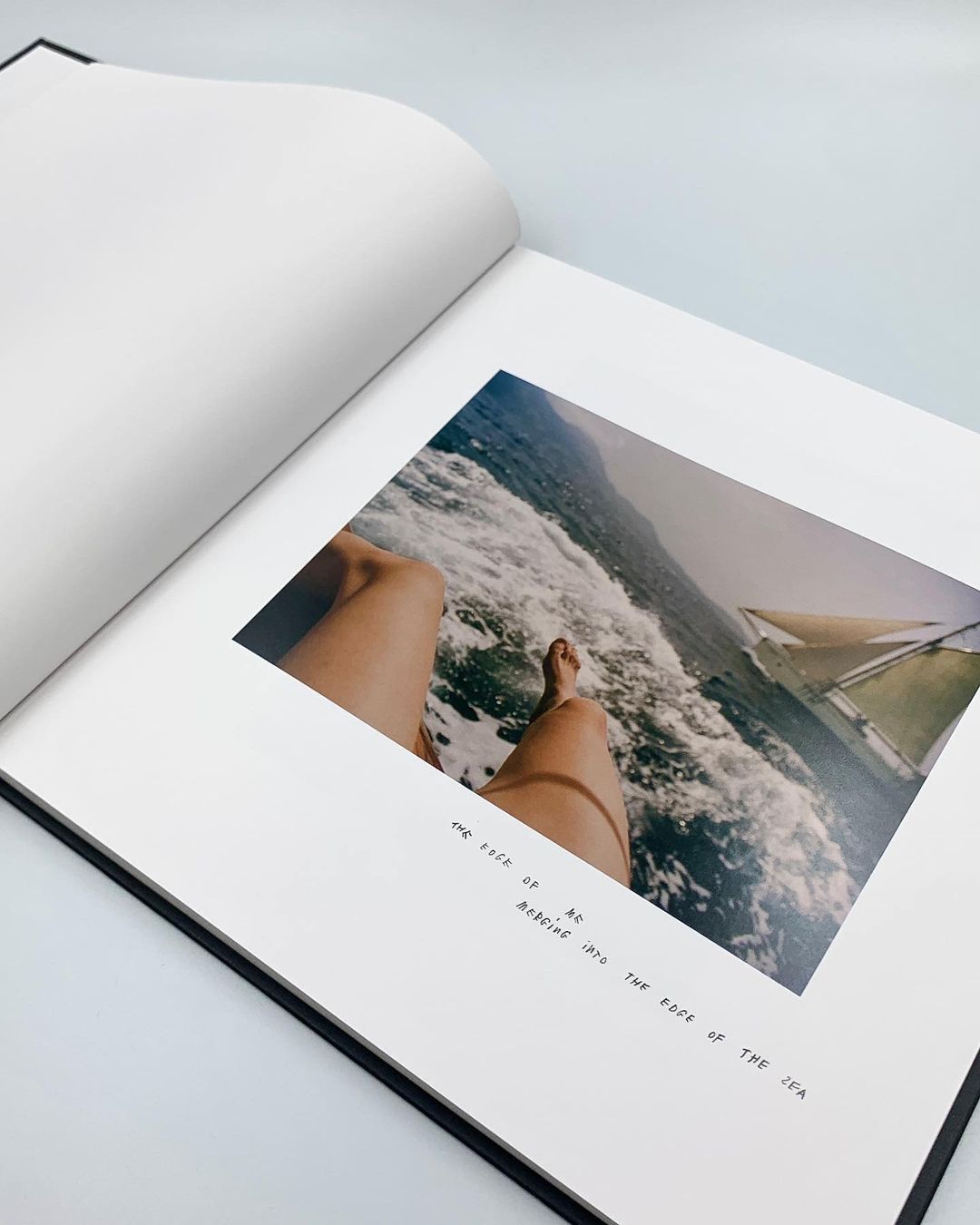 An open book features a photograph of legs on a boat amidst the sea, accompanied by black text on a white page in a minimalist style.