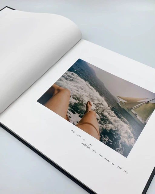 An open book features a photograph of legs on a boat amidst the sea, accompanied by black text on a white page in a minimalist style.