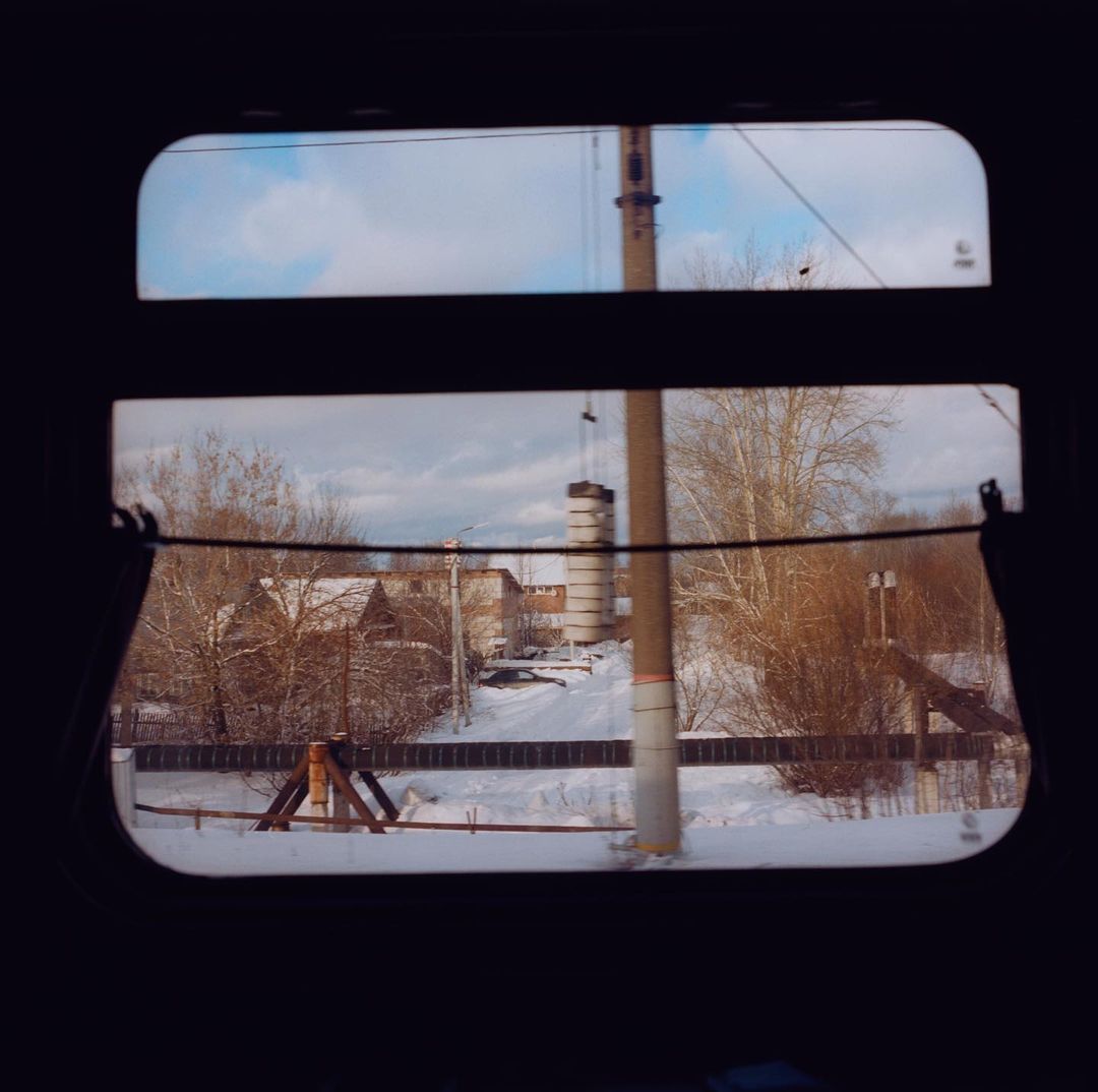 View from train window of snow-covered landscape with buildings, bare trees, and cloudy sky on winter day, capturing travel scenery.
