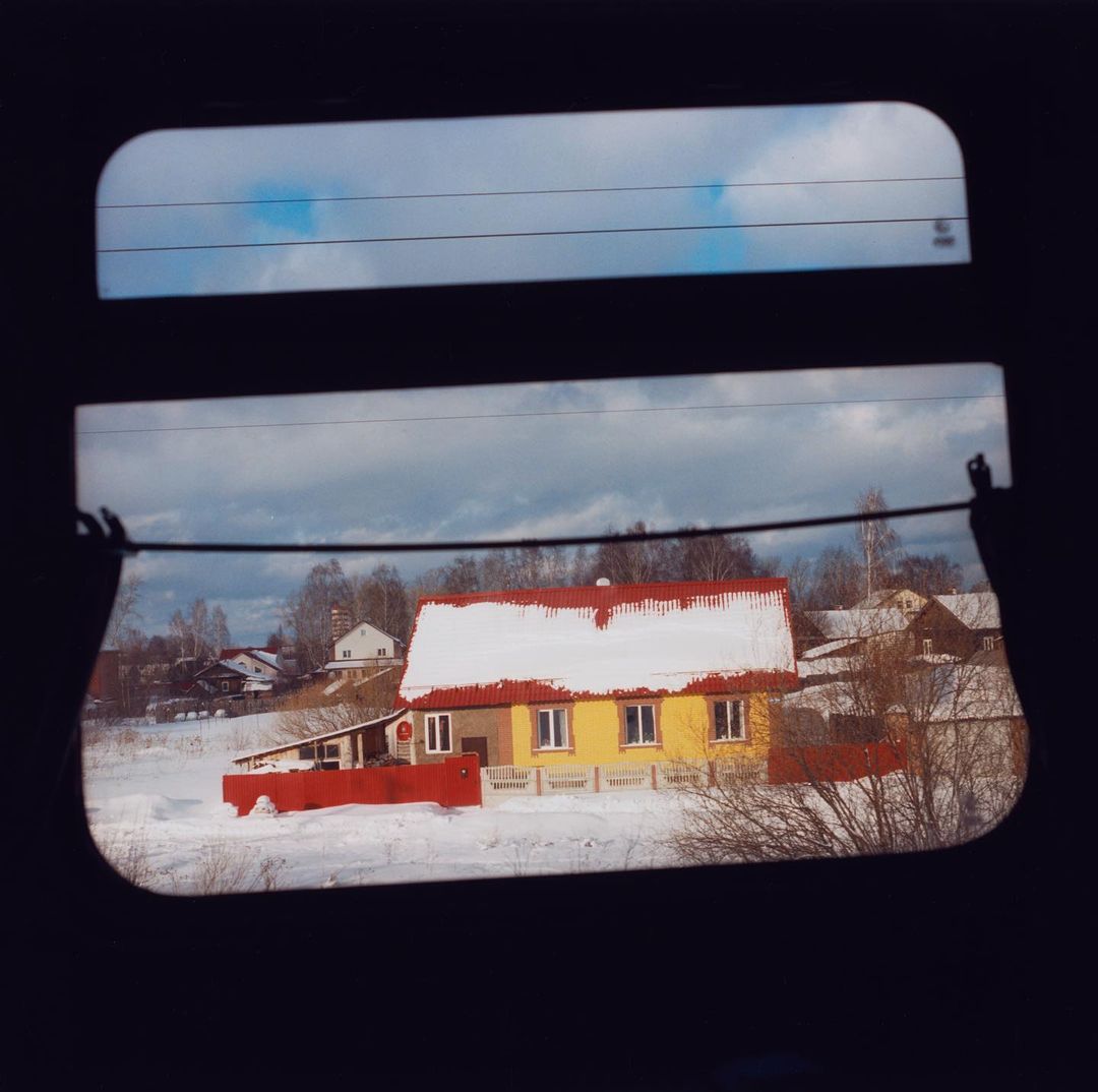 Snow-covered yellow house viewed from a train window, framed by black window edges, creating a wintery, abstract, and scenic view.