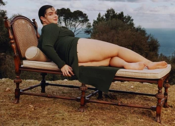 Woman in a green dress lying on a vintage wooden daybed outdoors with an ocean and sky background, photographed by Coco Capitan.