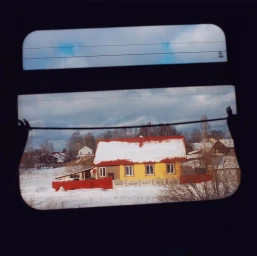 View through a train window showing a yellow snow-covered house against a cloudy sky by Coco Capitan