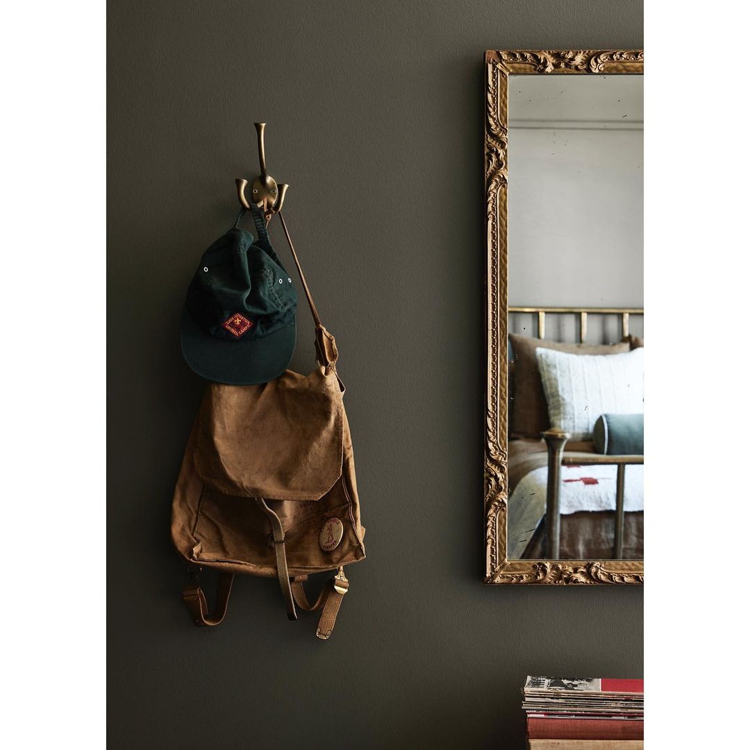 A brown backpack and green cap hang on a bronze hook on a dark green wall, next to a gold-framed mirror reflecting a bedroom interior.