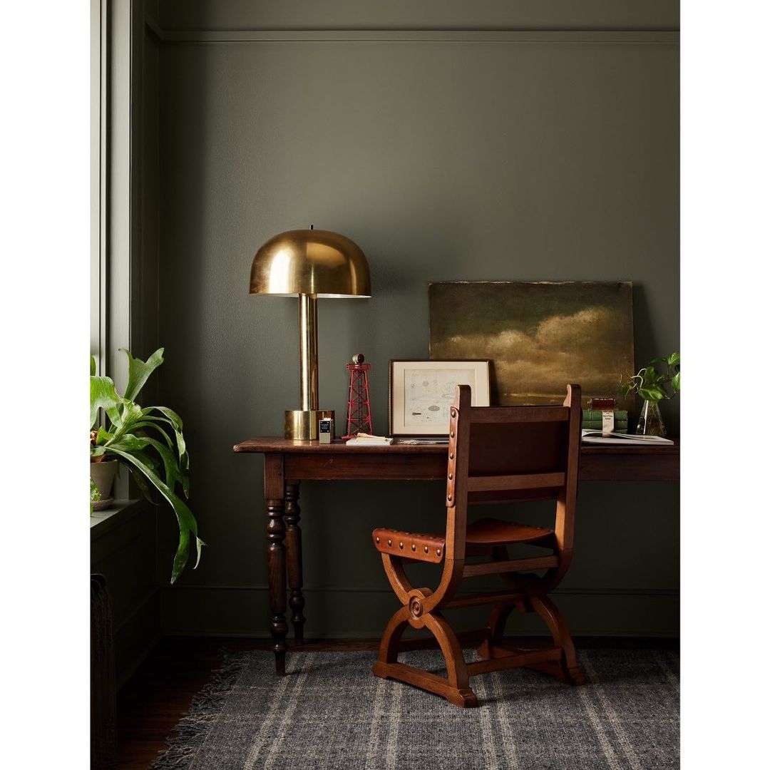 Interior view of a home office space featuring a wooden desk with a brass lamp, artwork, and a decorative chair on a striped rug.