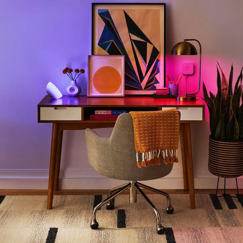 Desk setup featuring a mid-century modern desk with decor, including lamps, framed art, and a chair, set against a neutral wall.