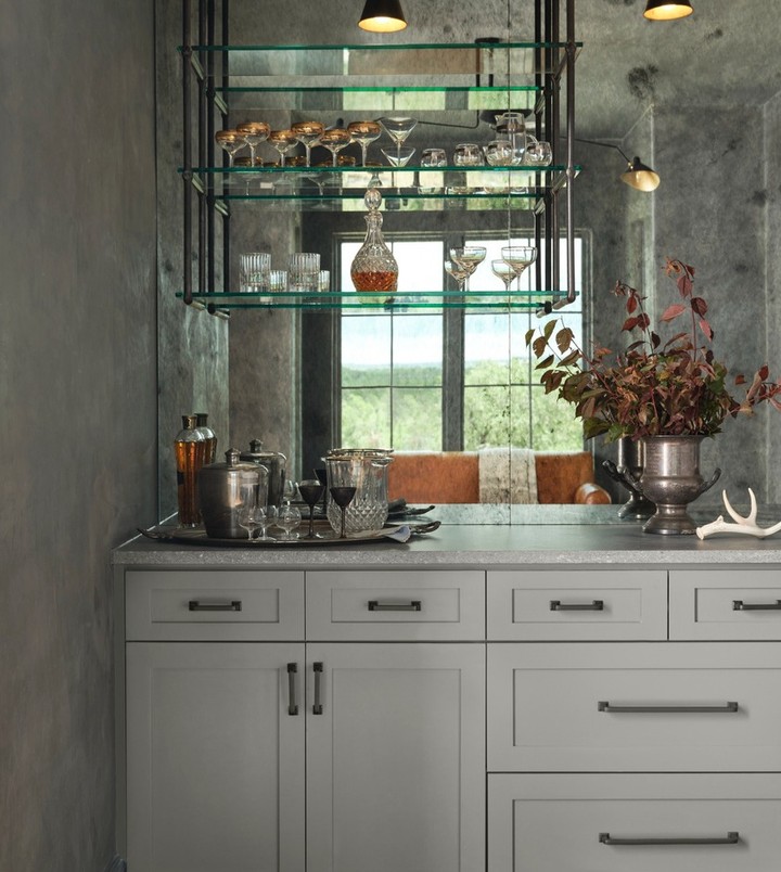 Well-stocked home bar with grey cabinets, open shelving, glass decanters and glasses, and silver serving pieces under warm lighting.