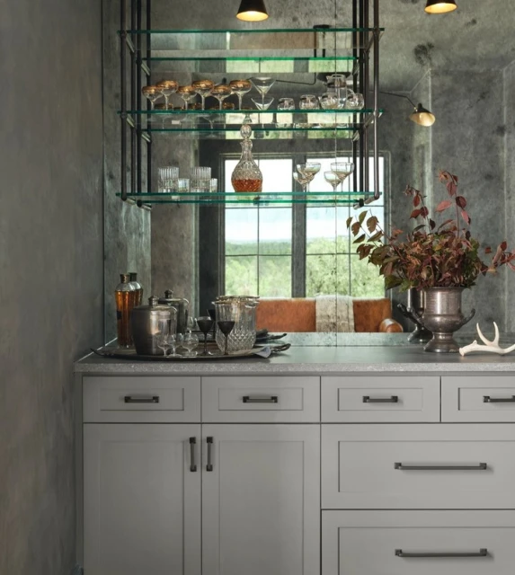 Well-stocked home bar with grey cabinets, open shelving, glass decanters and glasses, and silver serving pieces under warm lighting.