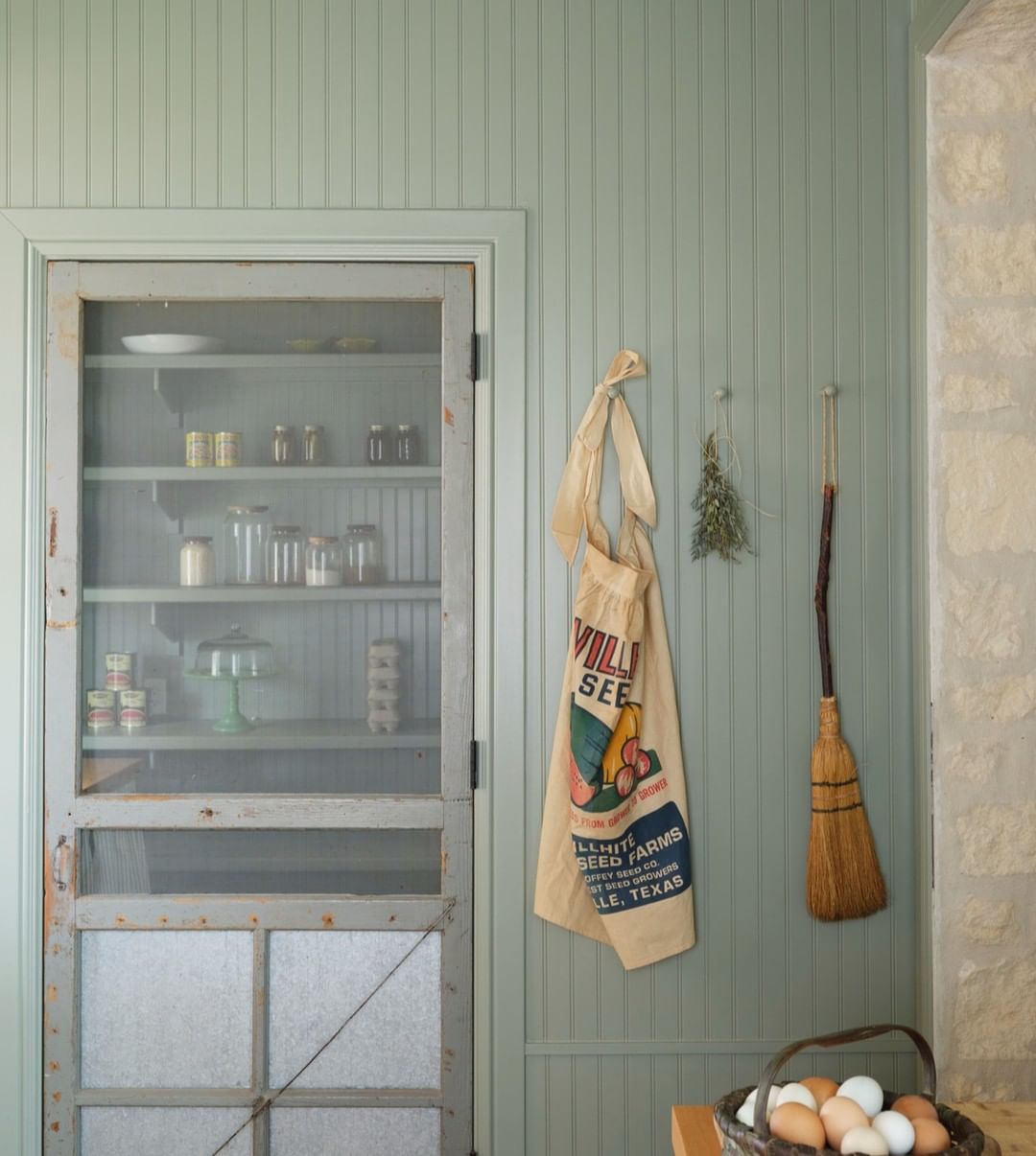 Interior view featuring a light green paneled wall, vintage cabinet with jars, a seed sack, dried herbs, and a broom.