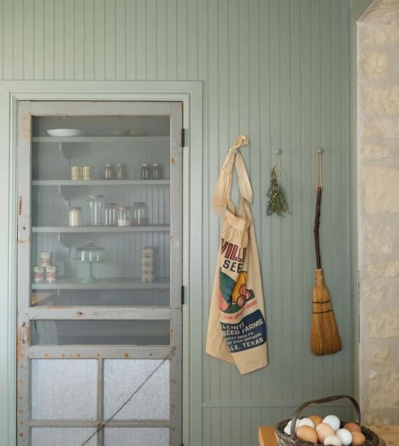 Interior view featuring a light green paneled wall, vintage cabinet with jars, a seed sack, dried herbs, and a broom.