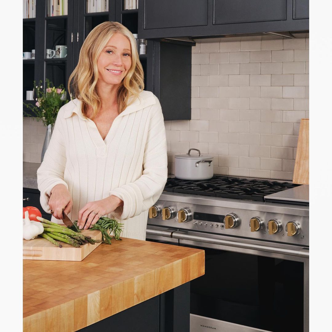 Smiling woman in a cream-colored sweater preparing asparagus on a wooden cutting board in a kitchen with stainless steel appliances.