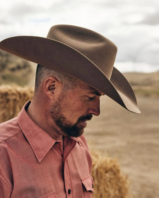 Man wearing a cowboy hat and a pink shirt in a rural outdoor setting with bales of hay and a cloudy sky in the background.