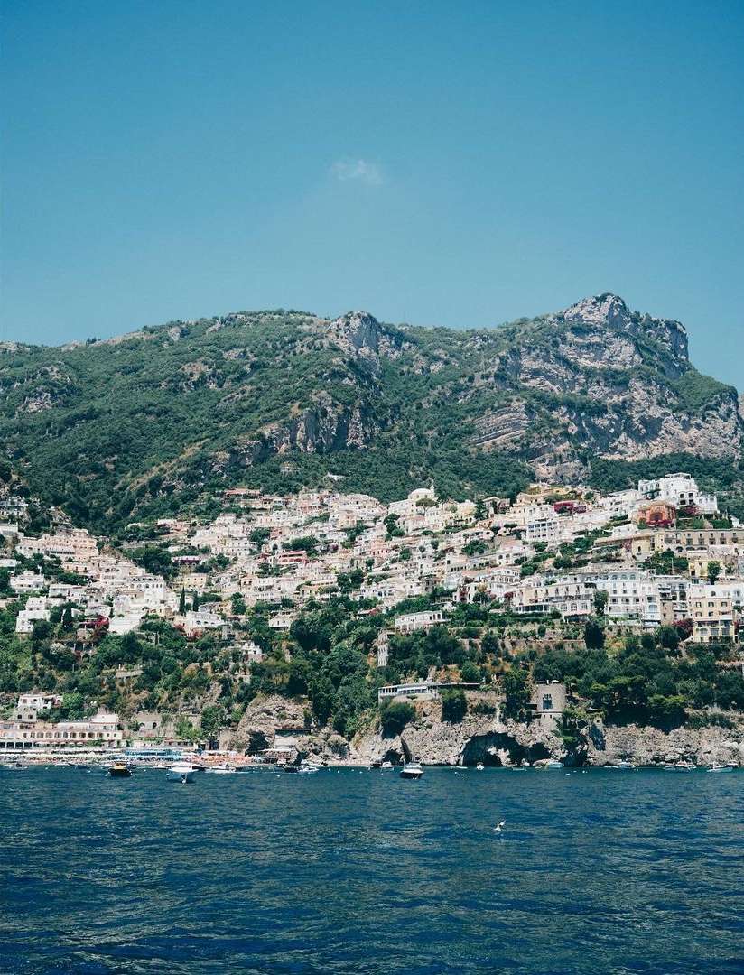 Picturesque view of the Amalfi Coast hillside with white buildings and green foliage, overlooking the blue sea under a clear sky.