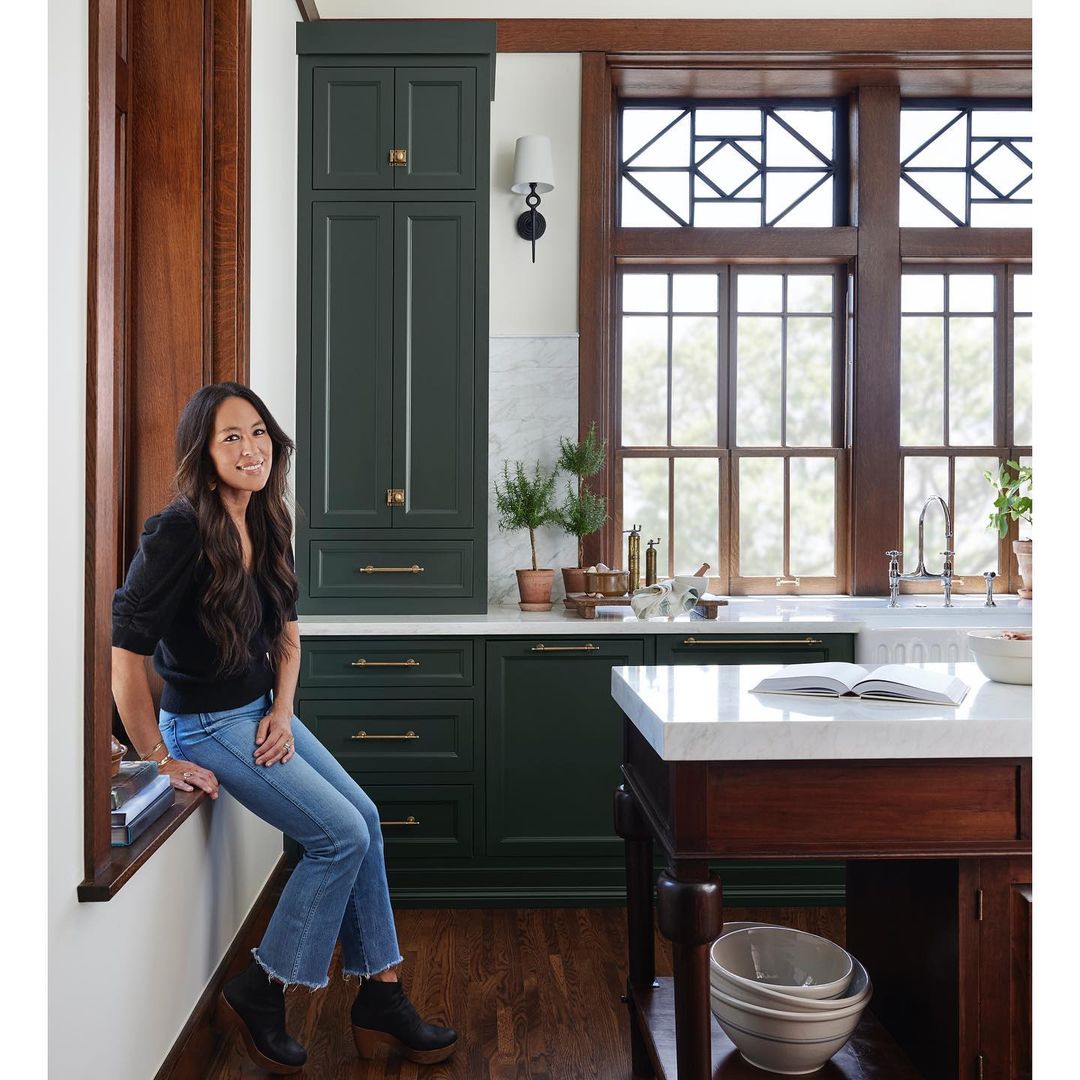 Woman sitting in a kitchen with dark green cabinets, light marble countertops, dark wooden floors, and large windows.