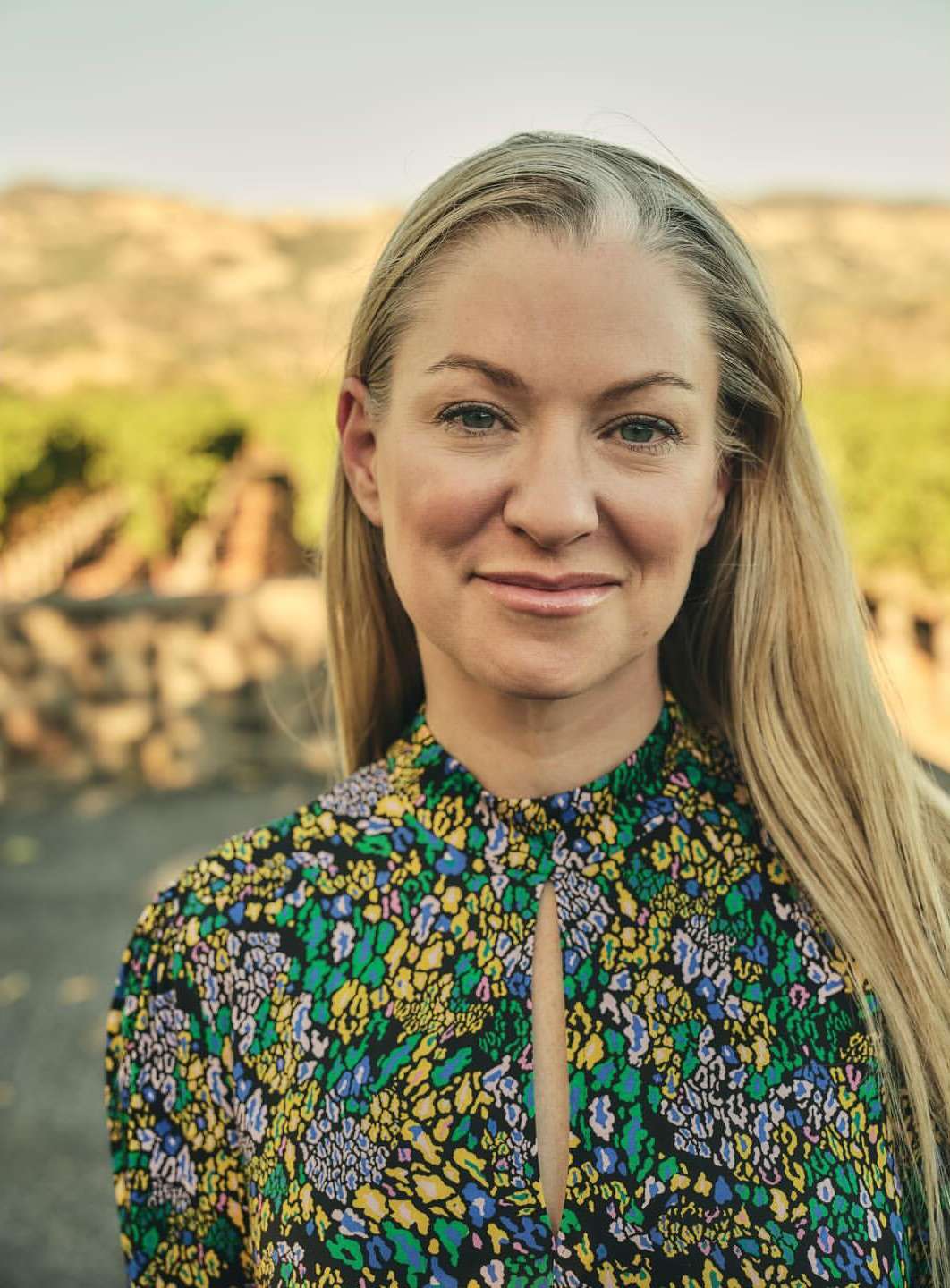 Close-up portrait of a blonde woman in a floral dress outdoors with a vineyard background, captured in natural lighting.