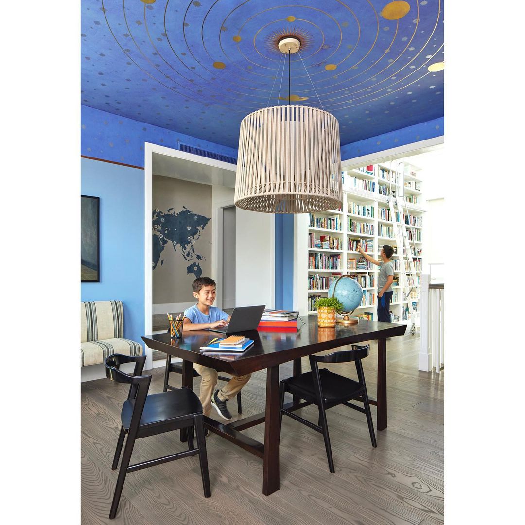 Young boy using a laptop at a dark wooden table in a blue-walled room with a ceiling mural of a solar system and hanging lamp.