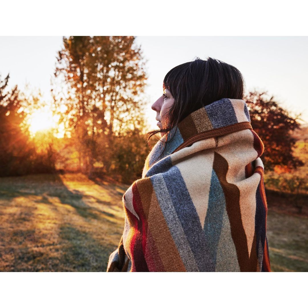 Woman wrapped in a patterned blanket gazes at the sunset in a serene outdoor setting with trees and golden light.