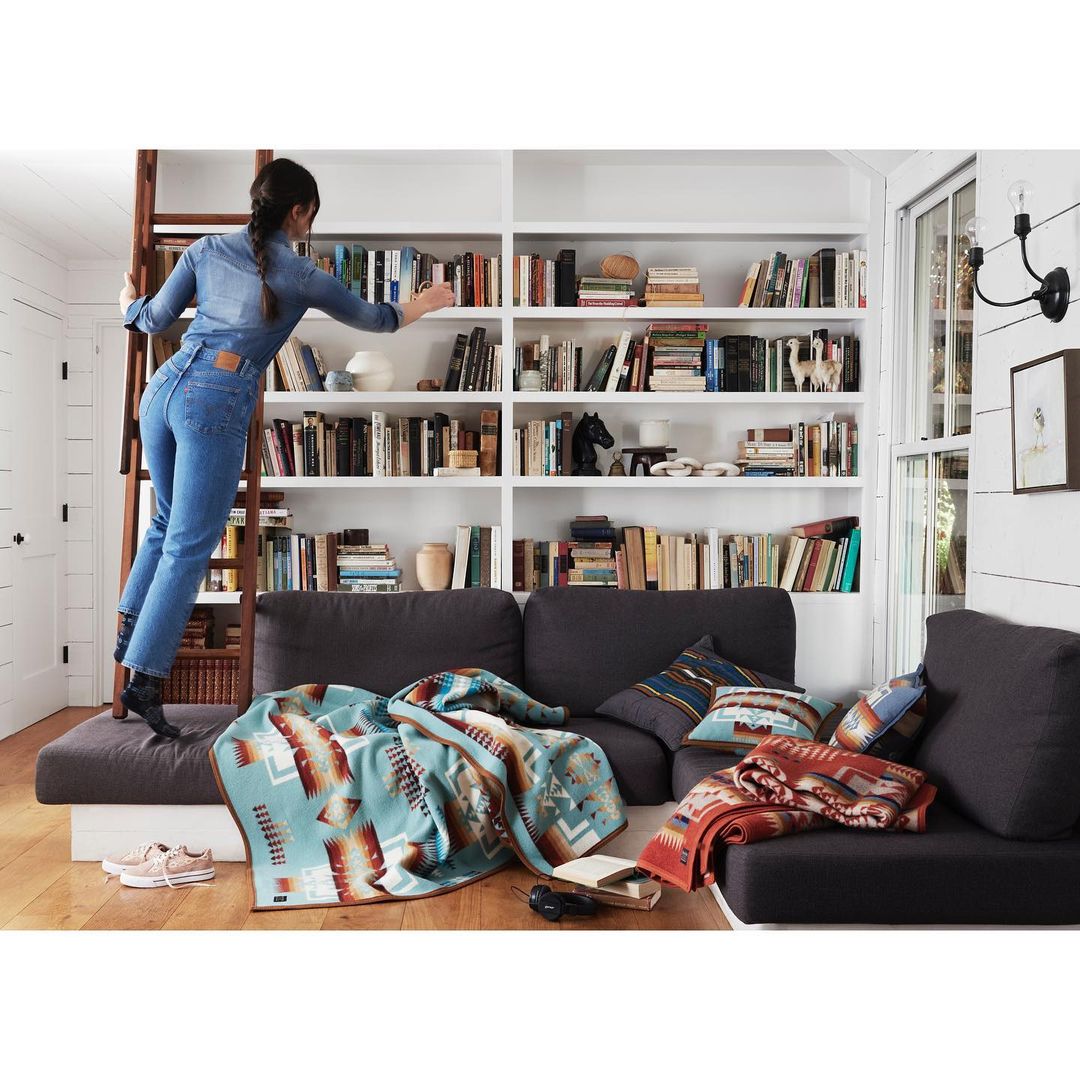 Woman in denim jumpsuit reaches for a book on a high shelf in a white bookshelf, with blankets draped over a grey sofa.