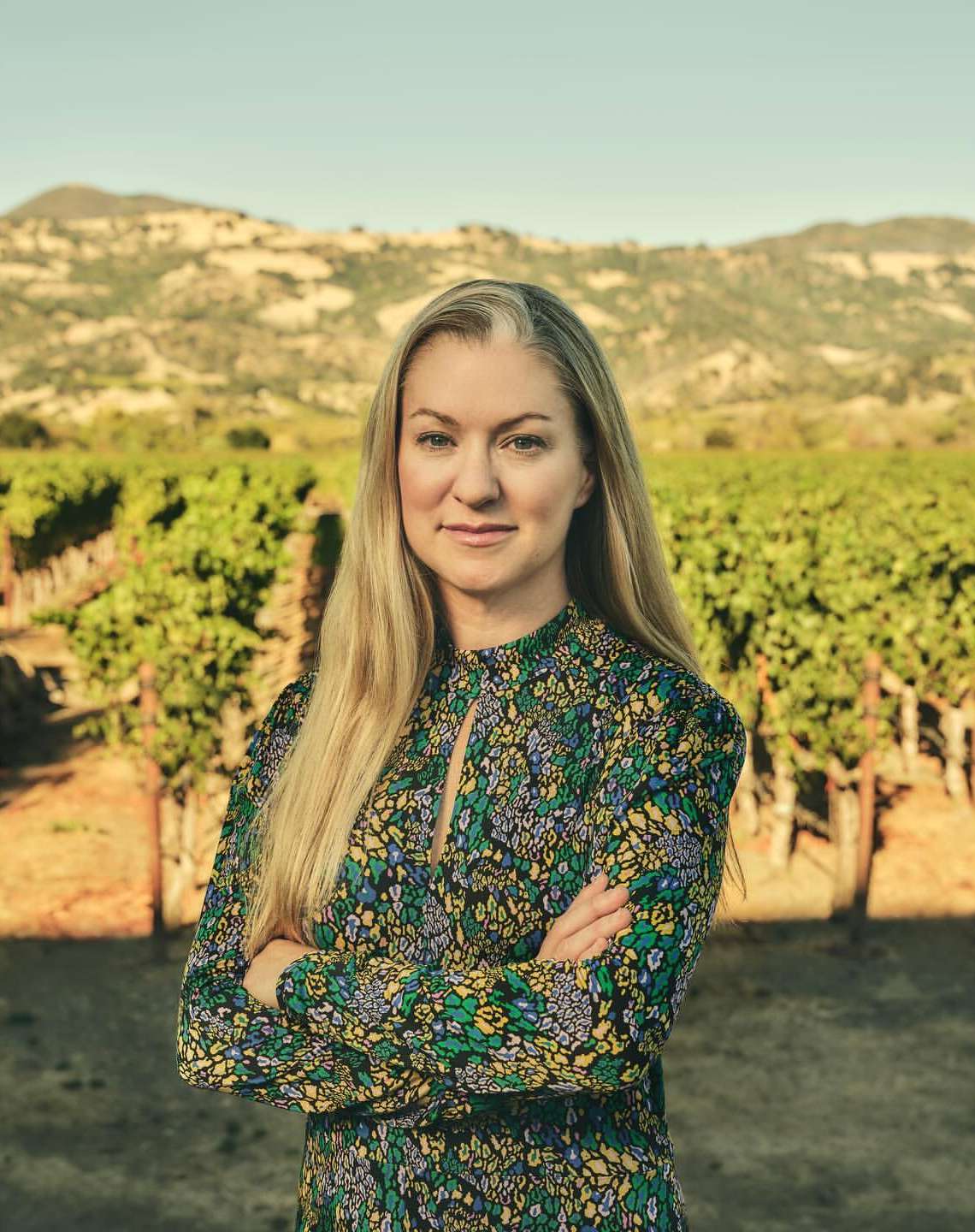 Portrait of a blonde adult female in a floral dress posing in a vineyard, with distant mountains visible under a blue sky.
