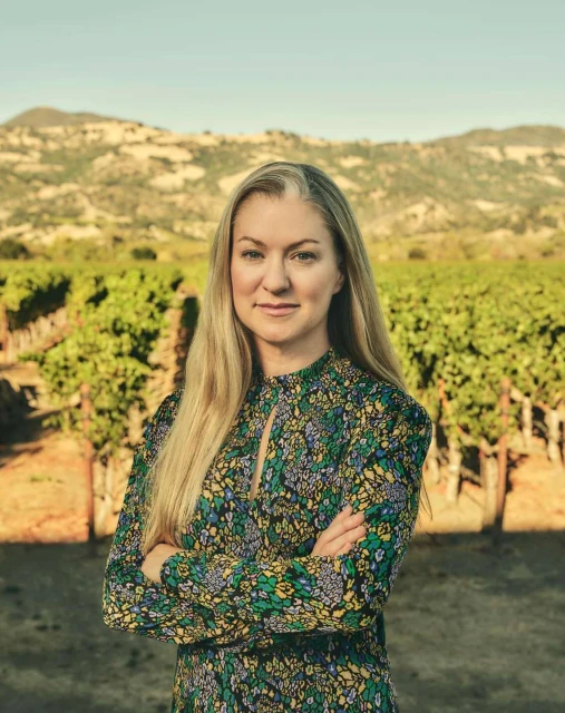 Portrait of a blonde adult female in a floral dress posing in a vineyard, with distant mountains visible under a blue sky.