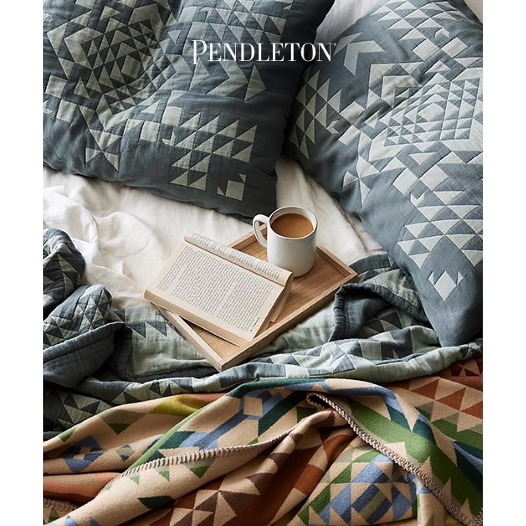 Still life featuring Pendleton blankets, pillows, a book, and a mug on a bed, showcasing a cozy and inviting bedroom interior.