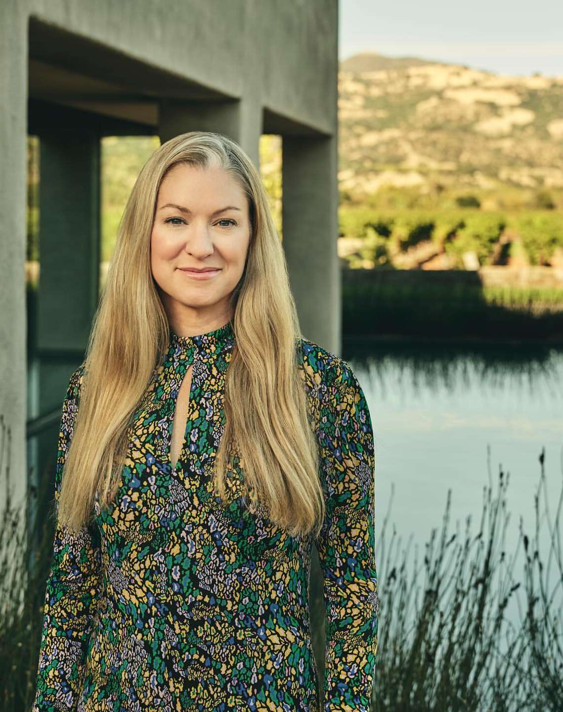 Portrait of a blonde woman wearing a patterned floral dress outdoors by a modern concrete structure and pond.