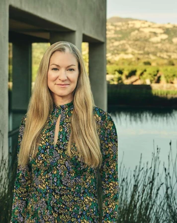 Portrait of a blonde woman wearing a patterned floral dress outdoors by a modern concrete structure and pond.