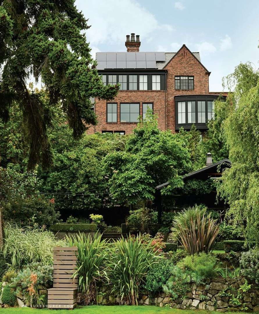 Exterior view of a large brick house with dark-framed windows and solar panels, surrounded by lush greenery and tiered gardens.