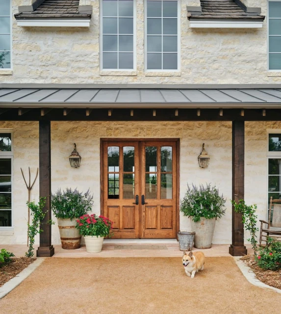 Exterior of a stone house with wooden doors, potted plants, a gravel path, and a corgi dog in the front yard.
