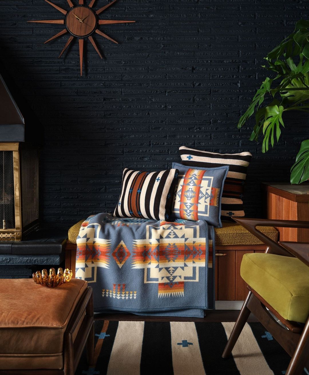 Interior scene features a patterned blanket, throw pillows, a wood frame chair, an ottoman, and a dark brick wall.