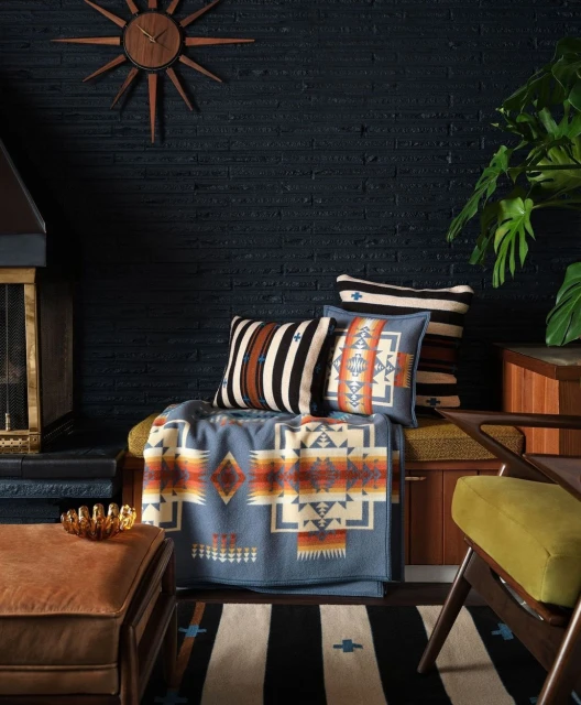 Interior scene features a patterned blanket, throw pillows, a wood frame chair, an ottoman, and a dark brick wall.
