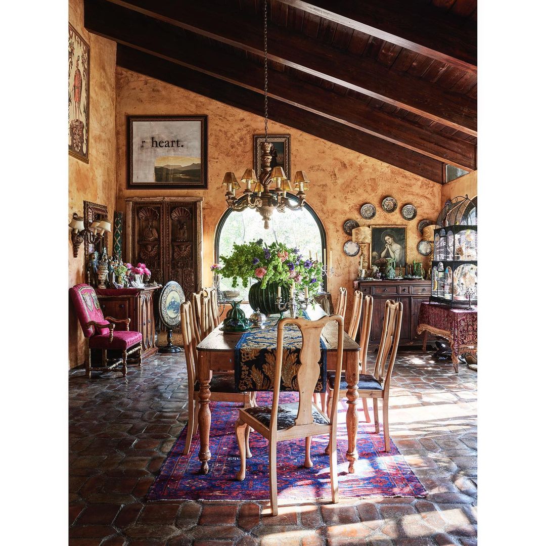 Warmly lit dining room with rustic wooden table, floral centerpiece, and mismatched chairs, featuring textured orange walls and tiled floors.