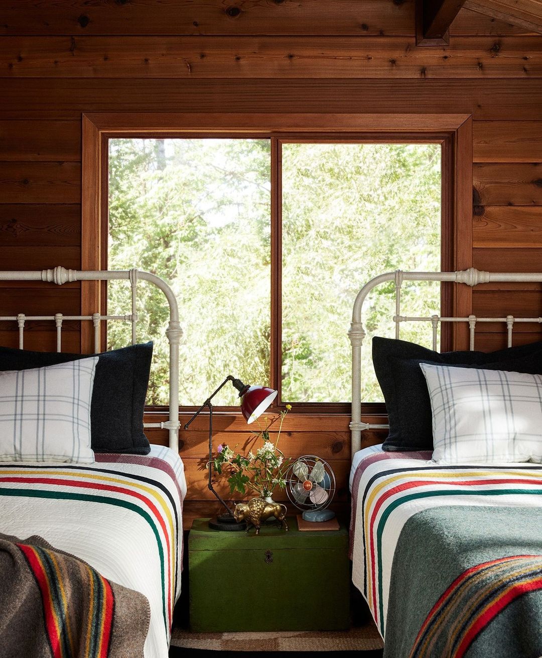 Cozy bedroom interior with two twin beds, striped blankets, throw pillows, a wooden nightstand, a table lamp, and a window with greenery.