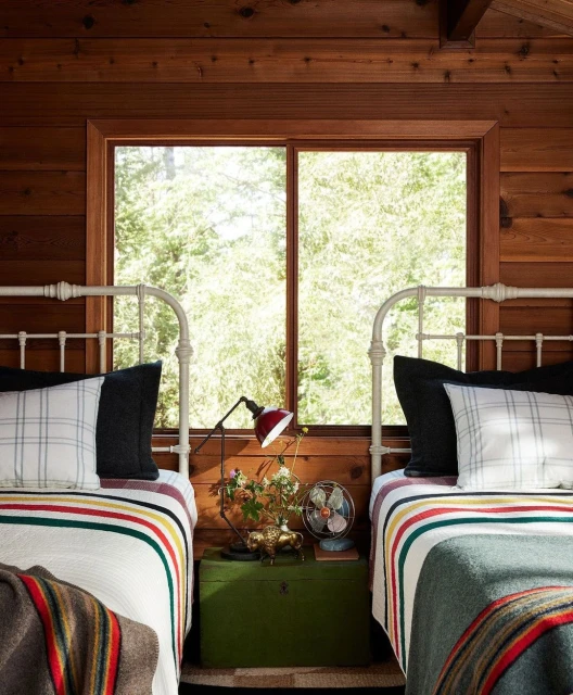 Cozy bedroom interior with two twin beds, striped blankets, throw pillows, a wooden nightstand, a table lamp, and a window with greenery.