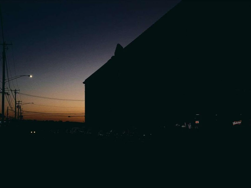 Silhouette of a dark building at dusk against a gradient sky with streetlights and power lines creating an urban evening scene.