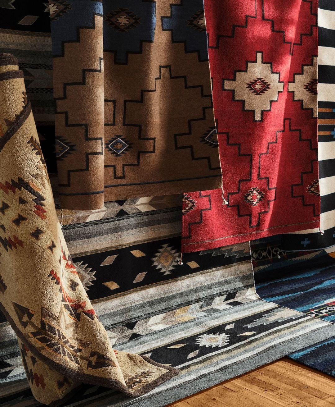 Display of various patterned, tan, brown, red, blue, and black wool rugs featuring geometric designs and rolled and flat layouts.