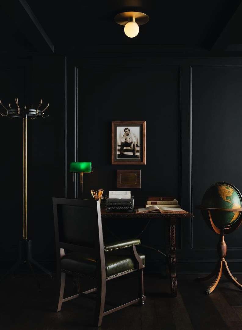 Dark interior with a writing desk set with a vintage typewriter, books, a lamp, and a globe, illuminated by an overhead light.