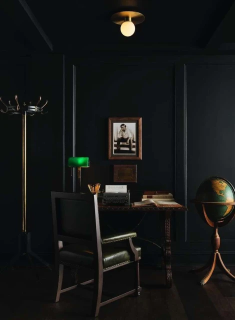 Dark interior with a writing desk set with a vintage typewriter, books, a lamp, and a globe, illuminated by an overhead light.
