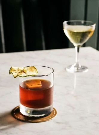 Two cocktails on a marble table: one brown liquid with apple garnish, one clear with a stem, set against a dark green background.