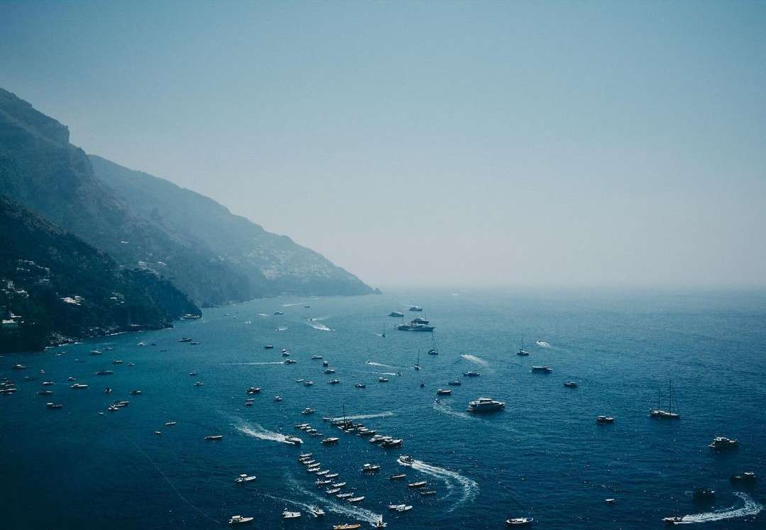 Scenic view of numerous boats on the blue ocean waters by a mountainous coast under a clear sky on a sunny day.