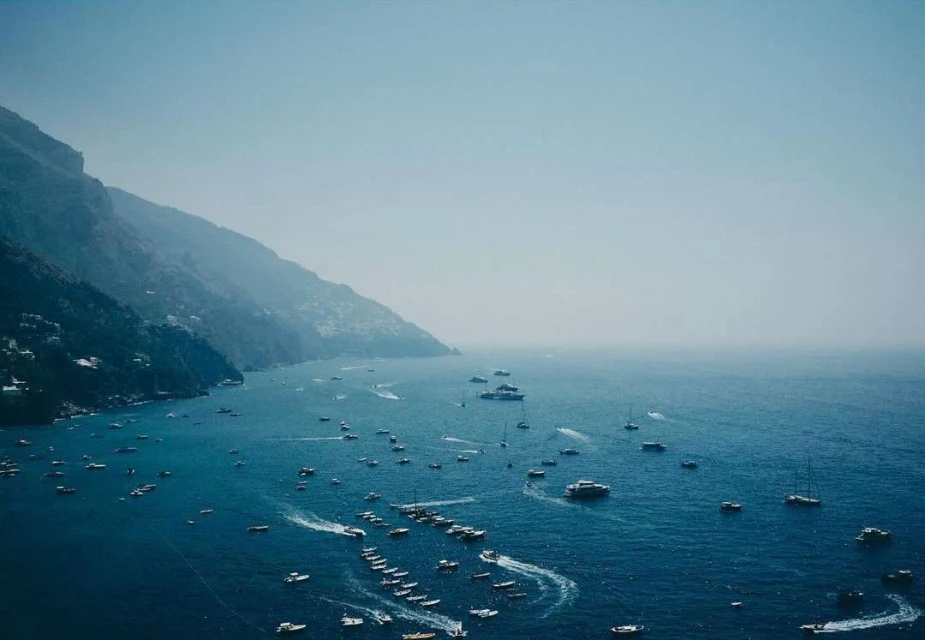 Scenic view of numerous boats on the blue ocean waters by a mountainous coast under a clear sky on a sunny day.