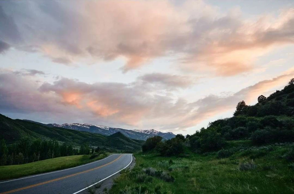 Scenic mountain road winds through lush green landscape under a pastel sunset sky with soft pink and white clouds.