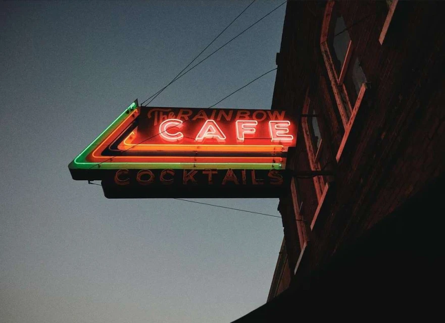 Neon sign for The Rainbow Cafe with cocktails is lit up against a dark building and evening sky, casting a warm glow.