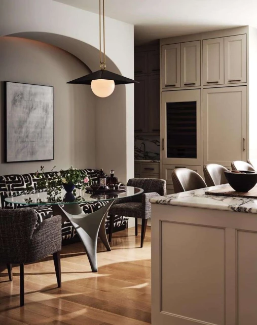 Contemporary dining area with glass table, patterned settee, and pendant lighting in a modern, neutral-toned home interior.