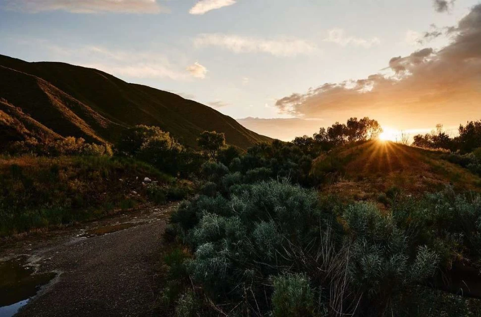 Scenic landscape of mountains and vegetation under a dynamic sky with the sun peaking through the clouds, creating a warm, golden light.