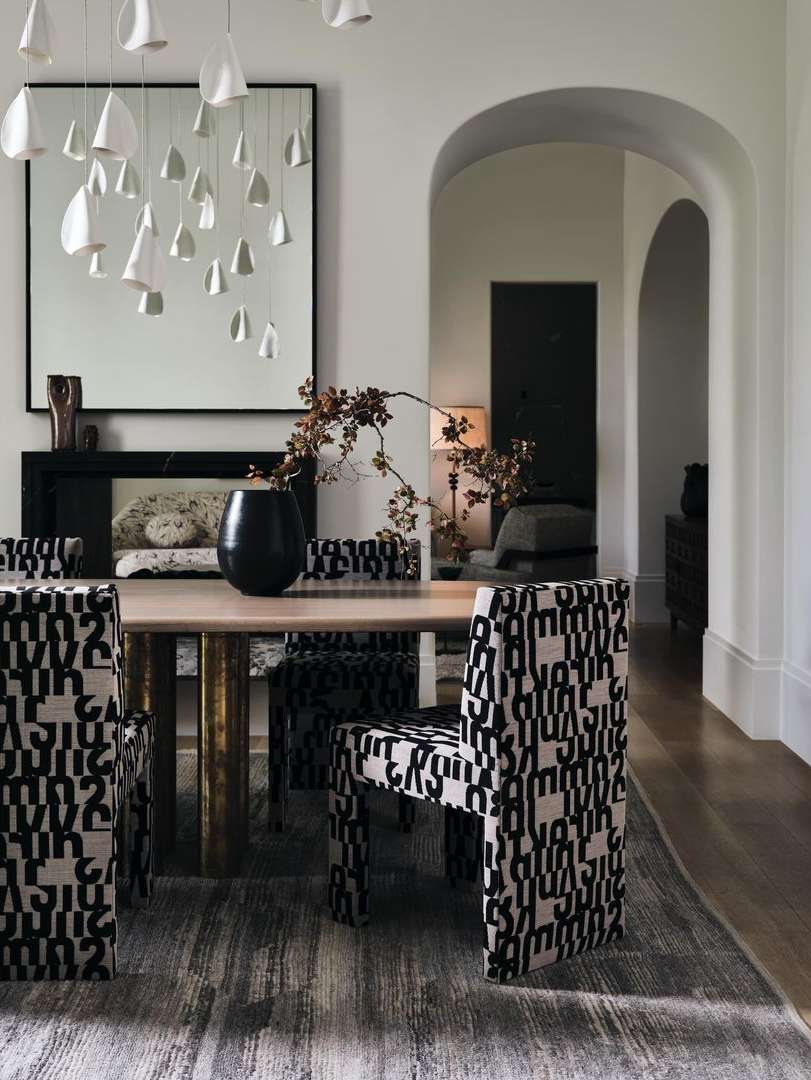 Dining room interior showcasing an oval wooden table with black vase, chairs with patterned upholstery, modern chandelier, and a fireplace.