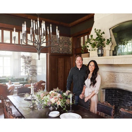 Man and woman standing together in a formal dining room with dark wood paneling, a large chandelier, and a stone fireplace.
