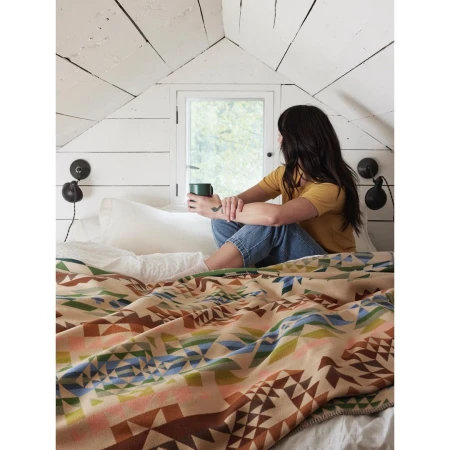 Woman sitting on a bed under a slanted white wood ceiling, holding a green mug and gazing out a small window.