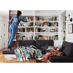 Woman in denim climbing a wooden ladder to reach books on white shelves in a living room.