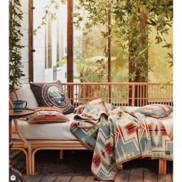 A rattan daybed draped with a patterned wool blanket and pillows inside a bright sunroom filled with hanging plants.