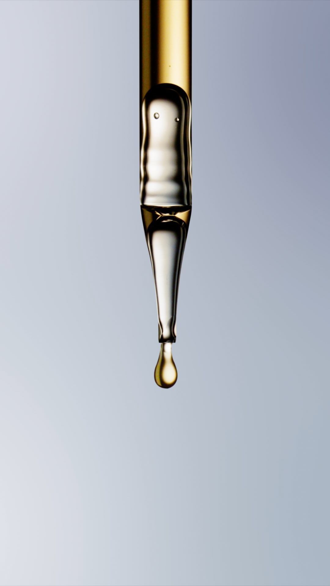 Close-up of a pipette filled with clear liquid with a yellow tint, poised to release a single drop against a light blue backdrop.